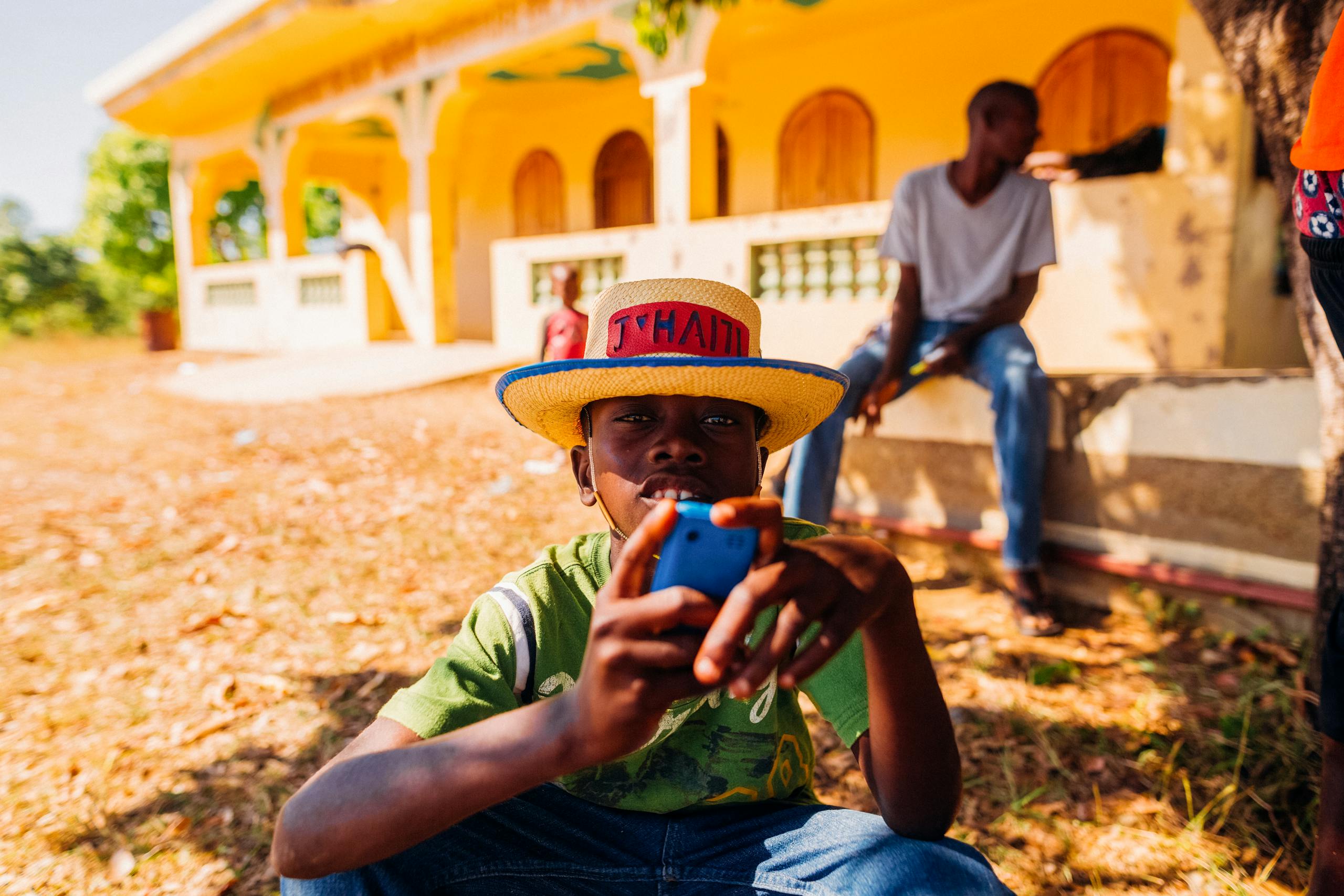 A young boy in a straw hat using a smartphone on a sunny day in rural Haiti.