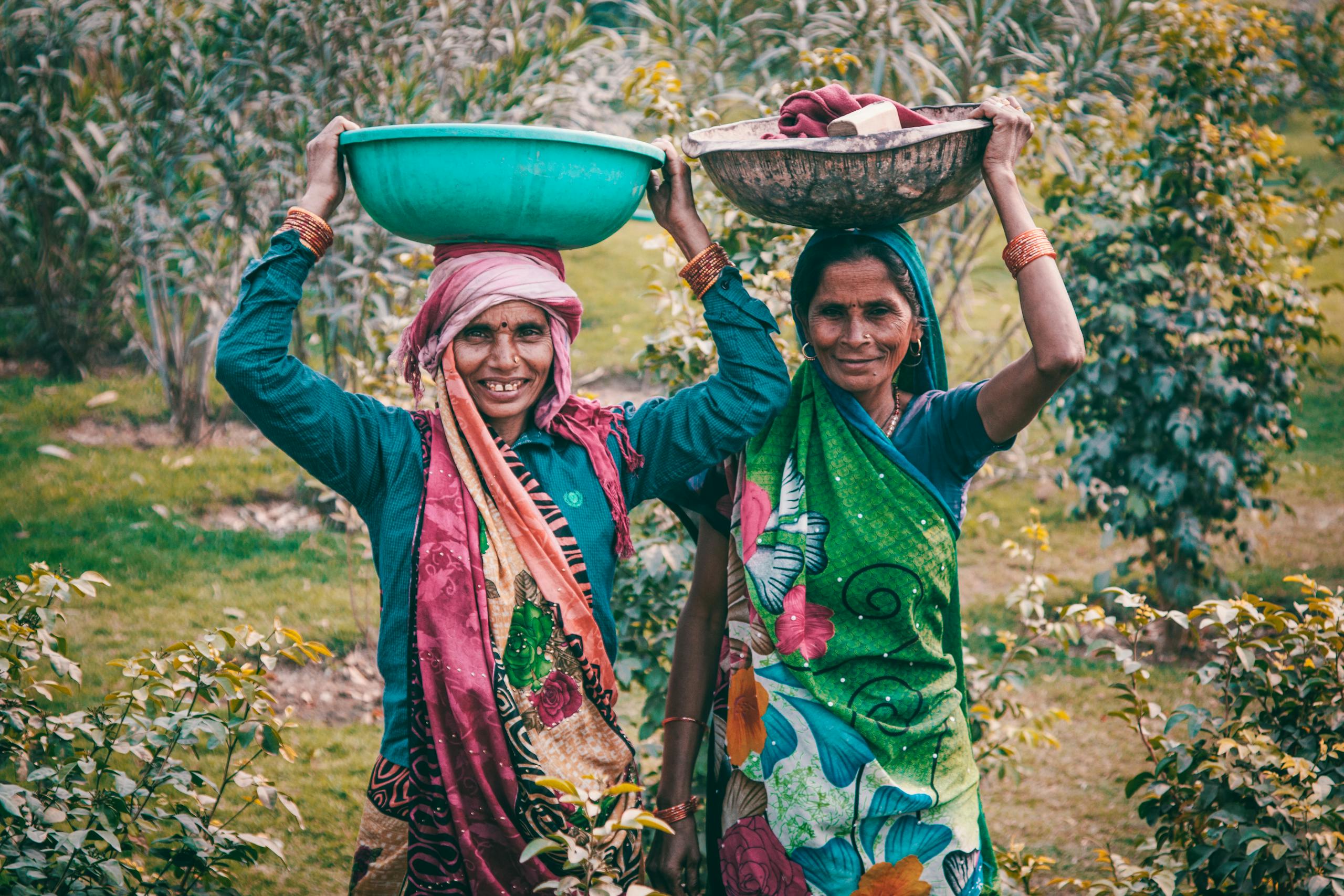 Two women wearing colorful traditional dresses smile outdoors, carrying baskets on their heads.